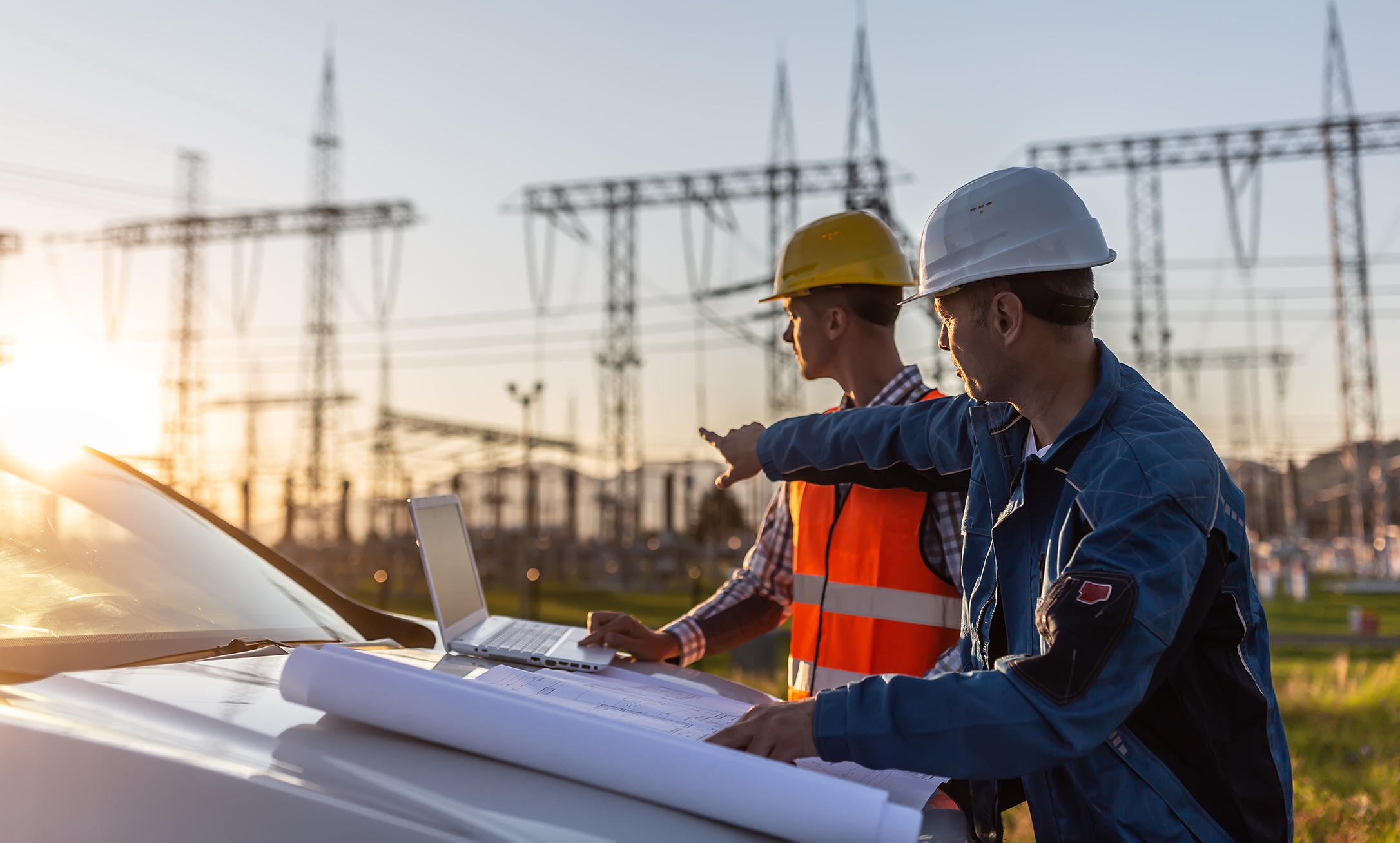 Two men with laptop on hood of truck pointing at power lines